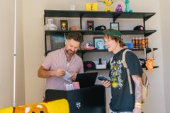 Two men standing in a room with shelves of toys and collectibles behind them, one holding a small item while looking at a laptop.