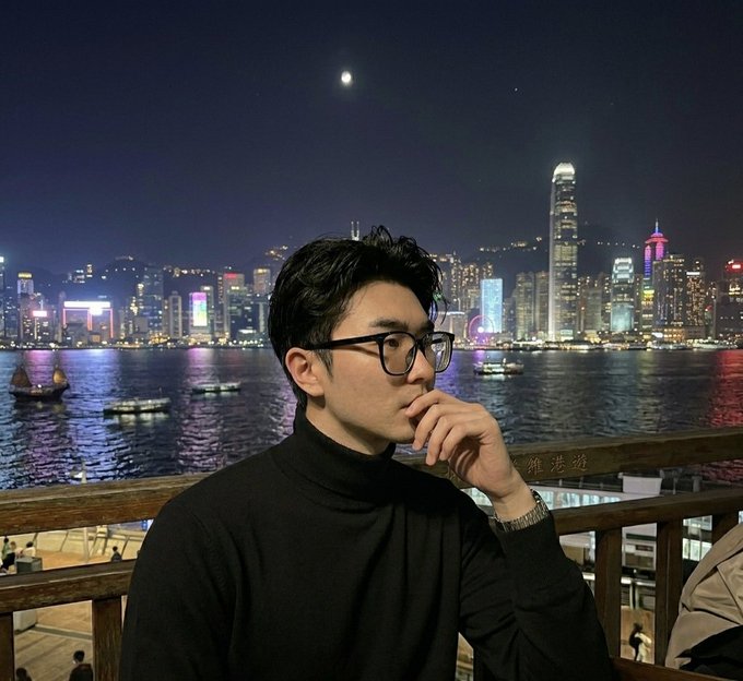 The image features a man wearing glasses, sitting at an outdoor table with his hand resting on his chin. He appears to be looking off into the distance or deep thought. The background of the photo shows a city skyline at night, which includes tall buildings that are lit up against the dark sky. There is also a boat visible in the water near the city.
