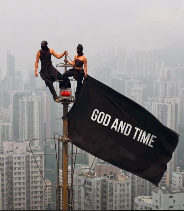 Dramatic photo of two masked figures standing atop a communications tower high above a hazy city skyline, holding a large black flag emblazoned with 'GOD AND TIME'.