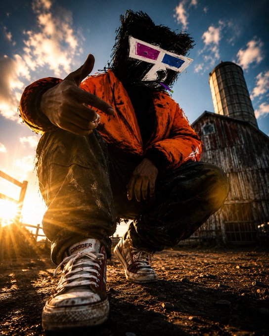 A low-angle photograph of a person crouching in mud wearing an orange jacket, ripped jeans, and Converse sneakers, their face obscured by a mask with 3D glasses, set against a sunset sky and rustic wooden buildings.