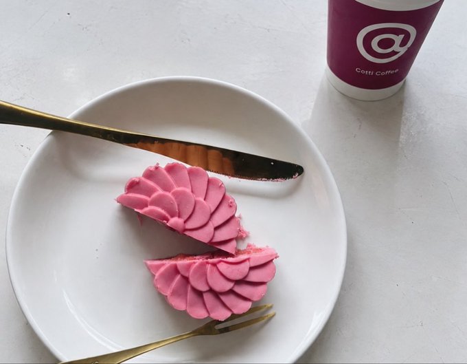 A pink, intricately patterned dessert cut in half sits on a white plate with gold utensils, next to a Cotti Coffee cup featuring its '@' logo.