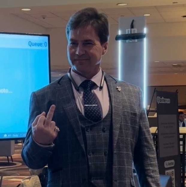 A man in a grey plaid three-piece suit and tie pointing his index finger upward in what appears to be a conference setting, with a screen showing "Queue: 0" in the background.