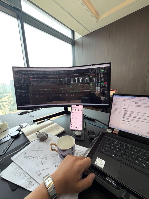 A trader works at a multi-screen setup displaying cryptocurrency charts, with a laptop showing "Universe" news on Bitcoin, while a handwritten note, coffee mug, and phone rest on the desk—reflecting the post's message about patiently accumulating BTC through market crashes and cycles.