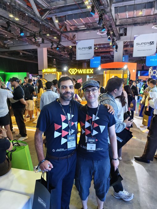 Two men wearing matching shirts with a triangular pattern pose together at what appears to be a conference or expo, with "WEEX" and "MEXC" signage visible in the background.