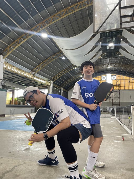 Two young men in pickleball gear pose playfully on an indoor court, each holding a black paddle.