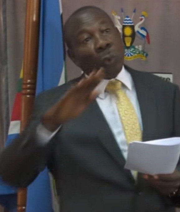A man in a dark suit and yellow tie speaks animatedly while holding papers, with the Ugandan national flag and coat of arms visible behind him. The post "Salary immediately it enters" suggests he is making a statement about salary payments, fitting the impression of a government official addressing the topic publicly.