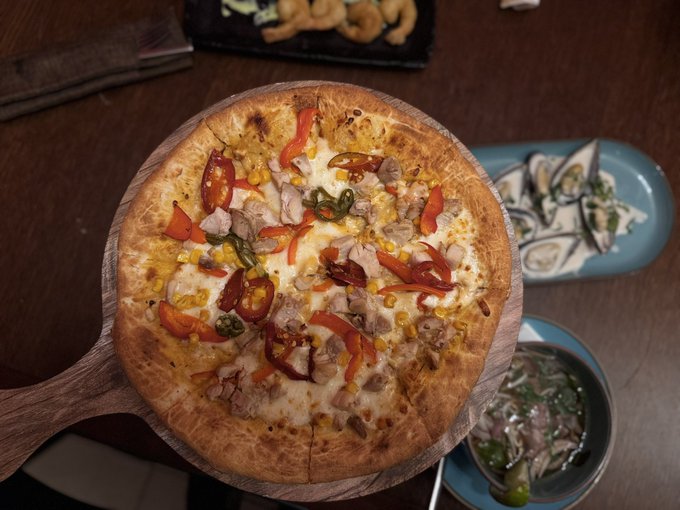 An overhead view of a pizza topped with chicken, red peppers, corn, and jalapeños, served on a wooden board alongside side dishes.