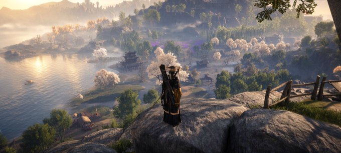 A scenic video game screenshot showing a character with a katana standing on a rocky cliff overlooking a peaceful village, lake, and flowering trees in the distance.
