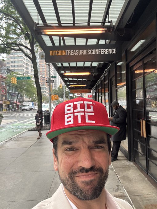 A smiling man takes a selfie wearing a red cap with "BTC BTC" printed on it, standing outside a venue displaying a "BITCOINTREASURIESCONFERENCE" sign on the marquee. The image captures the casual, celebratory vibe of "We out here," highlighting his presence at a Bitcoin-focused event in the city.