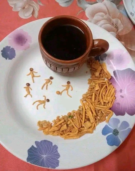 A terracotta mug of dark coffee or tea on a floral-patterned paper plate, accompanied by cereal sticks and small figurine-shaped biscuits depicting people playing soccer.