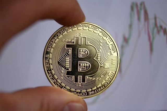 A close-up photo of fingers holding a physical Bitcoin coin with a candlestick chart visible in the background, symbolizing cryptocurrency trading.