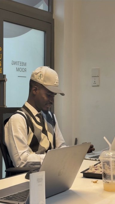A man wearing a white cap and argyle vest sits working on a silver Apple MacBook at a table, with a partially visible 'Meeting Room' door behind him.