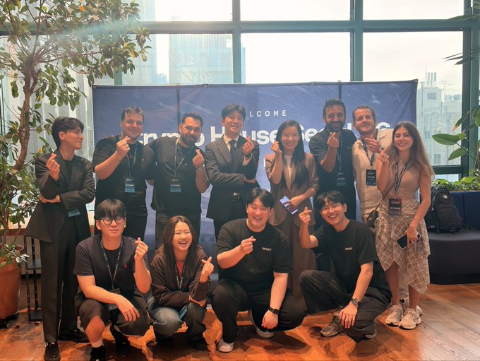 A group photo of thirteen people posing in front of a blue 'WELCOME HOUSE OF NG' banner, with several individuals making finger-heart gestures.