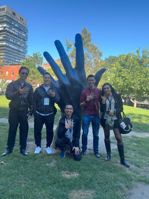 A group of five people posing in front of a large sculpted hand sculpture in a park setting, with some holding up peace signs.