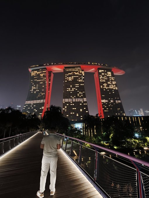 A person in white clothing stands on a lit bridge at night, gazing up at the iconic Marina Bay Sands hotel with its three illuminated towers and rooftop SkyPark.