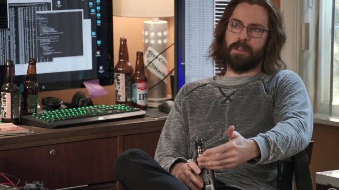 A bearded man with long hair and glasses wearing a gray shirt sits in an office chair, gesturing with his hand while holding a beer bottle, with a desk behind him displaying a computer monitor, keyboard, and several beer bottles.