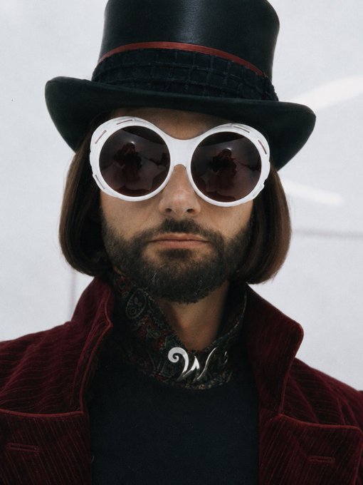 A man with shoulder-length brown hair, a beard, and large round white sunglasses wearing a black top hat and a red corduroy jacket, with a paisley scarf around his neck.