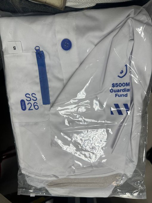 A sealed plastic package containing a white shirt/smock with a blue zipper, labeled 'S' (small) and 'SS 26 3X40,' along with a '$500M Guardian Fund' logo and a stylized 'M' emblem.