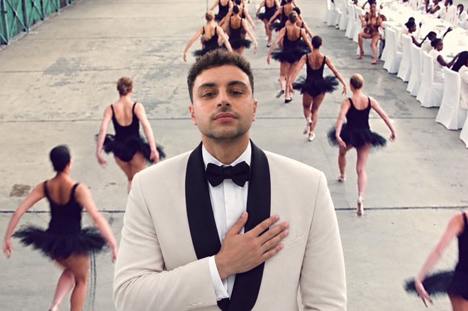 A man in a white tuxedo with a black bow tie stands in the foreground with his hand on his chest, while ballerinas in black tutus dance away from the camera in the background.