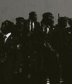 A silhouetted group of figures stands in shadow, with some appearing to hold raised weapons or tools against a light gray background.