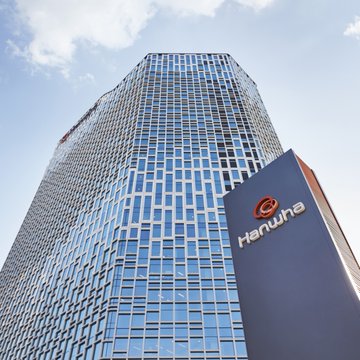 A low-angle shot captures a tall modern skyscraper with a glass facade, featuring a 'Hanwha' logo sign on a lower section against a partly cloudy sky.
