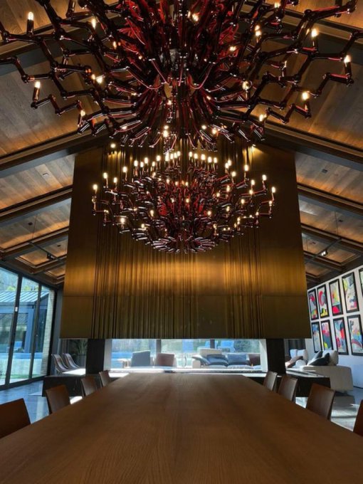 An opulent room features a massive wooden conference table beneath two striking red and black chandeliers, with large gilded curtains and a wall of framed artwork in the background. The luxurious real estate setting visually anchors the post's message, contrasting traditional property income with crypto's "fake yield" to emphasize why tokenizing real-world assets like real estate brings tangible value onchain.