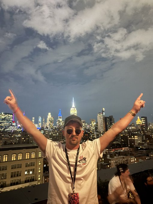 A man wearing sunglasses and a cap points both index fingers toward the New York City skyline at dusk, including the Empire State Building.