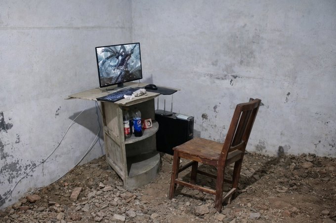 A makeshift computer setup in a ruined concrete room, with a monitor displaying an action scene on a small wooden stand next to a weathered wooden chair on a rubble-covered floor.