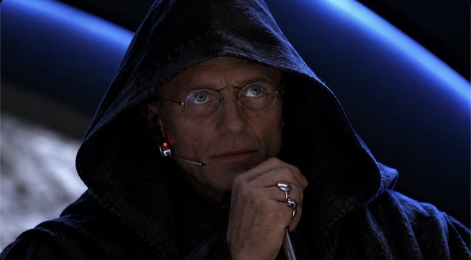 A hooded figure with round glasses and a headset, resembling Emperor Palpatine, gazes thoughtfully while holding a cane with a silver ring on his finger.