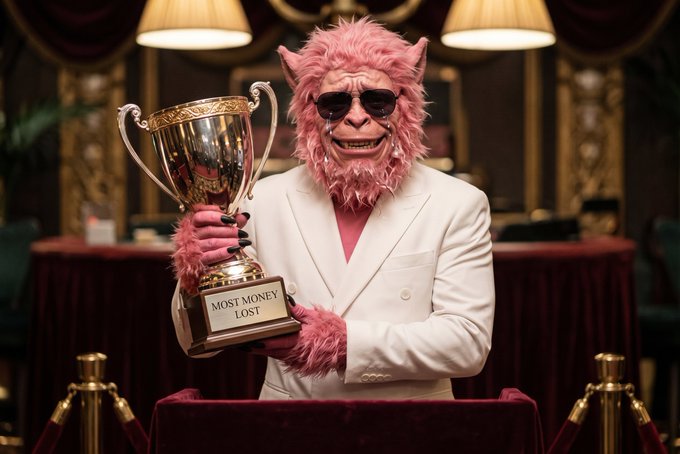 A pink monkey character wearing a white suit and sunglasses holds a gold trophy that reads 'MOST MONEY LOST' in a formal setting with velvet ropes.
