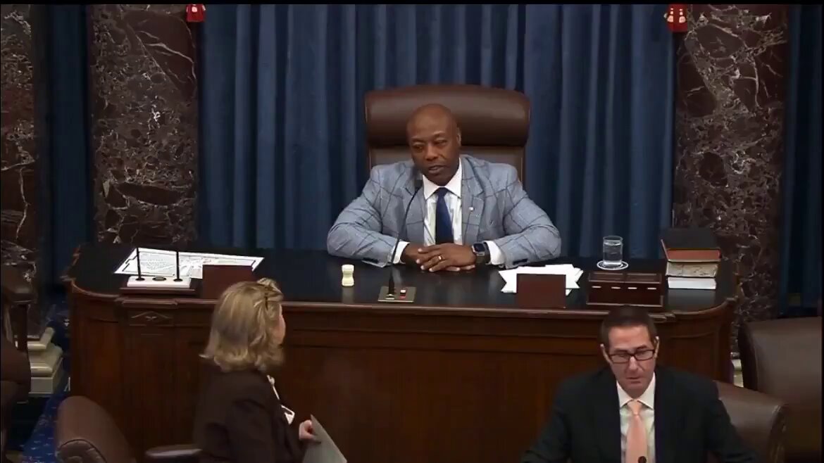 The image shows a man sitting at a desk, possibly in a courtroom or a similar setting, with two other people present - one person seated behind him and another person standing next to him. The scene appears to be related to a legal proceeding or an important decision being made. The focus of the image is on the man's position at the desk, which suggests that he might be involved in a case or a significant event taking place within the courtroom.