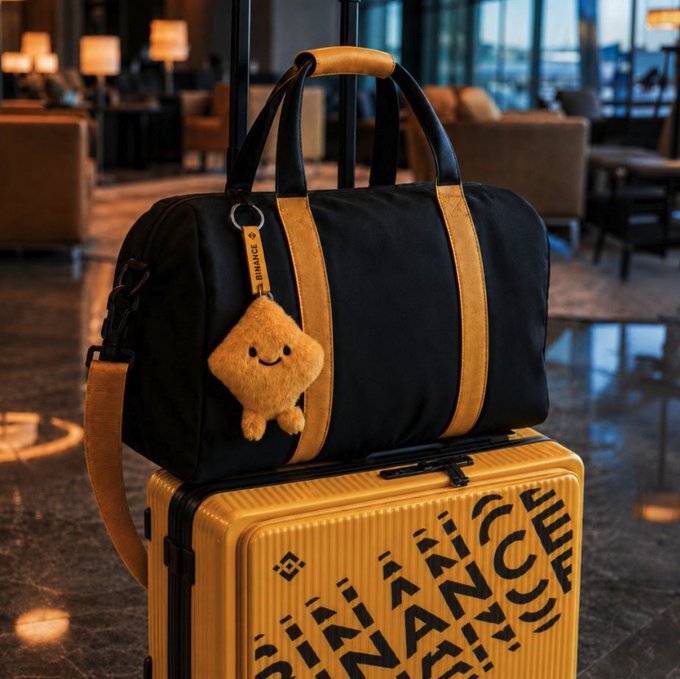 A black and gold Binance duffel bag rests atop a matching yellow suitcase branded with bold "BINANCE" lettering, accompanied by a plush Binance mascot tag, fitting the post's playful nod to crypto-themed travel upgrades.