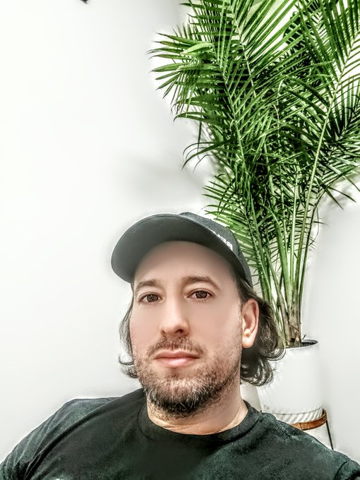 A bearded man wearing a black baseball cap and t-shirt poses for a selfie next to a potted palm plant.