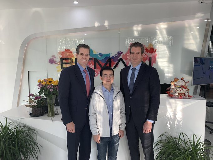 Three men pose together in an office lobby; two are dressed in dark business suits with ties, and the man in the middle wears a light grey jacket and blue shirt, standing in front of a white desk with a colorful 'BINANCE' logo on the glass wall behind them.