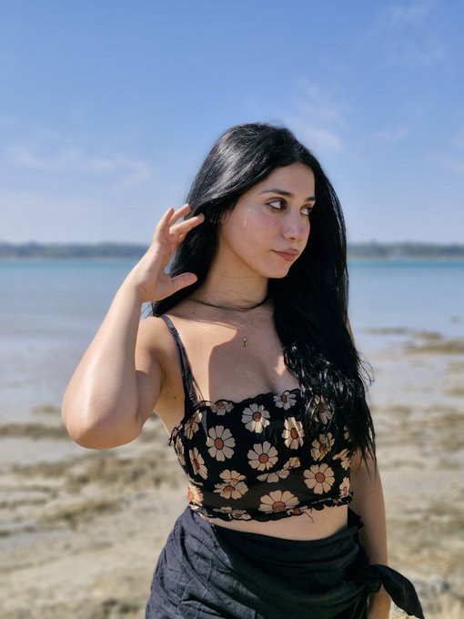 A young woman with long dark hair stands on a beach wearing a black floral crop top and dark skirt, posing with one hand near her hair against a backdrop of ocean and sky.