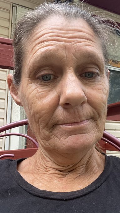A close-up selfie-style portrait of an older woman with gray hair pulled back, visible wrinkles, and blue eyes, standing in front of a house with red siding.