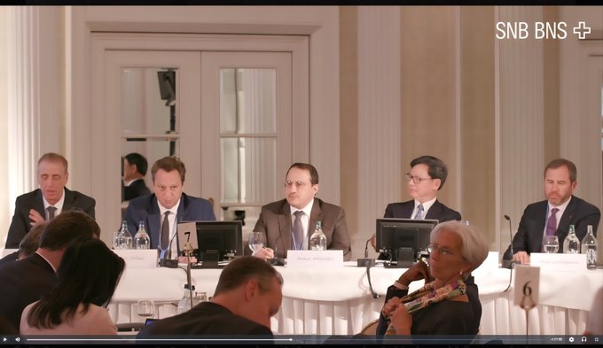 A formal panel discussion features several seated men in business attire (including a "7" name placard and a "6" placard) with name plates including "Jeremy Allix" visible, while the "SNB BNS" logo appears in the top right corner. The image relates to the post's context of a high-level international finance meeting, showing attendees at what appears to be an official Swiss National Bank/Bank for International Settlements event, with audience members like the woman in the foreground possibly representing figures such as Christine Lagarde referenced in the caption.