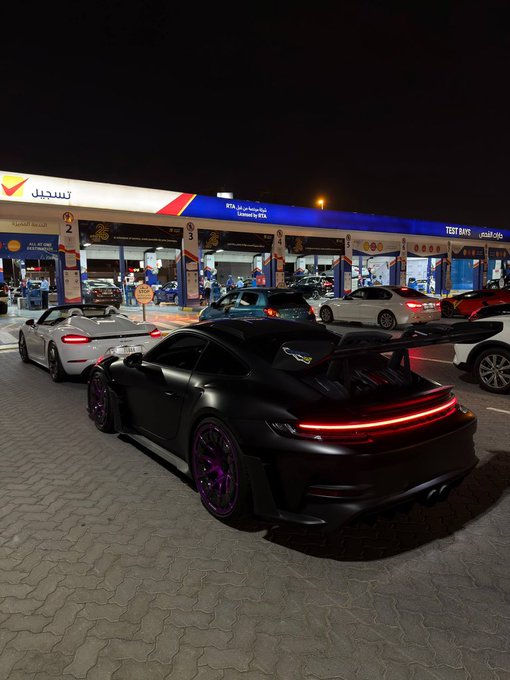 A matte black Porsche 911 GT3 with purple wheels and a large rear wing is parked outside an ADNOC gas station ("Clinic #14, TEST BAYS 2-6") at night, flanked by other sports cars. The image embodies the post's message about cashing in gains to buy tangible rewards—like a dream car for yourself or loved ones—rather than watching paper profits evaporate in a round trip.