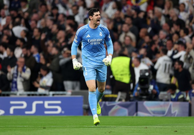 A Real Madrid goalkeeper in a light blue kit with "Emirates FLY BETTER" sponsorship celebrates on the field at the Santiago Bernabéu stadium with a PS5 advertisement visible on the pitch-side boards.