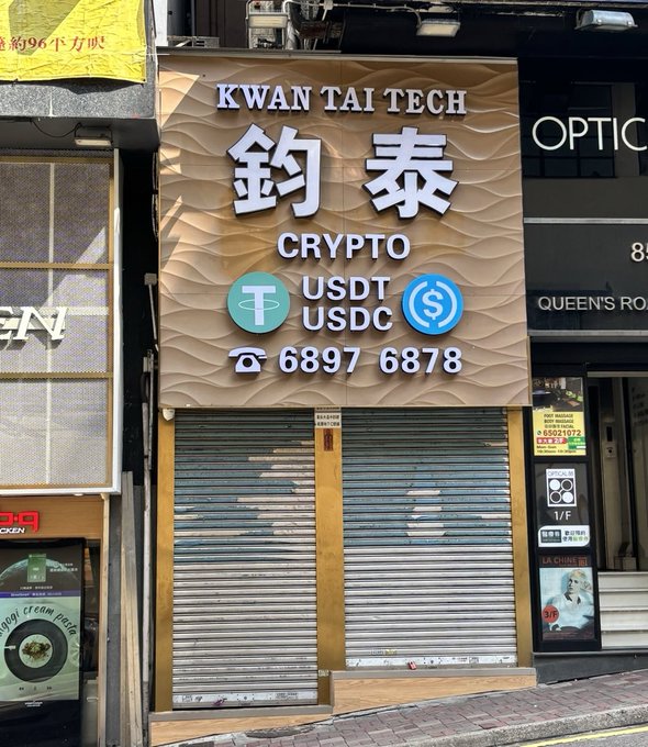 A storefront sign reads 'KWAN TAI TECH 鈎泰 CRYPTO USDT USDC ☏ 6897 6878' above a closed metal shutter on what appears to be Queen's Road.