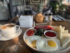 A classic breakfast spread featuring fried eggs, toast, and condiments sits on a wooden table alongside a latte and pineapple. The scene captures an ordinary, unhurried morning—contrasting ironically with the post's claim about the "boring" crypto lifestyle, since even the most exciting industries begin with simple routines.