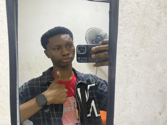 A young man takes a mirror selfie with his phone, wearing a plaid jacket over a red shirt and a smartwatch, giving a thumbs up.
