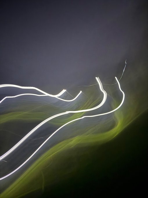 An abstract long-exposure image features wispy white light trails curving across a dark background with green, smoke-like wisps.