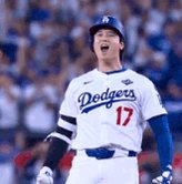 The image features a baseball player wearing a Dodgers jersey with the number 17, standing on a field during a game. The player appears to be celebrating or reacting to something happening on the field, possibly related to their performance or an exciting moment in the game. The background shows other players and spectators, indicating that this is a professional baseball game.