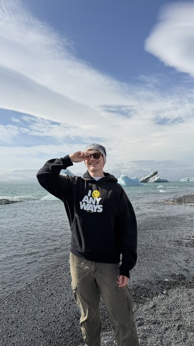A person stands on a rocky beach in front of the ocean, shielding their eyes from the sun while wearing a black hoodie that reads 'IT IS WHAT IT IS ANY WAYS' with a smiley face, along with sunglasses and a bandana.