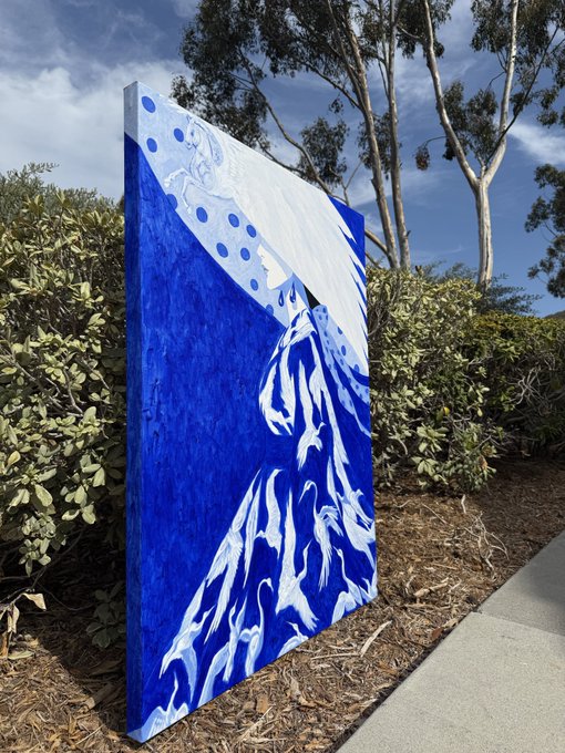 A large, freestanding outdoor canvas sits on mulch near a sidewalk, featuring a blue and white painting of a figure with wings or flowing fabric.