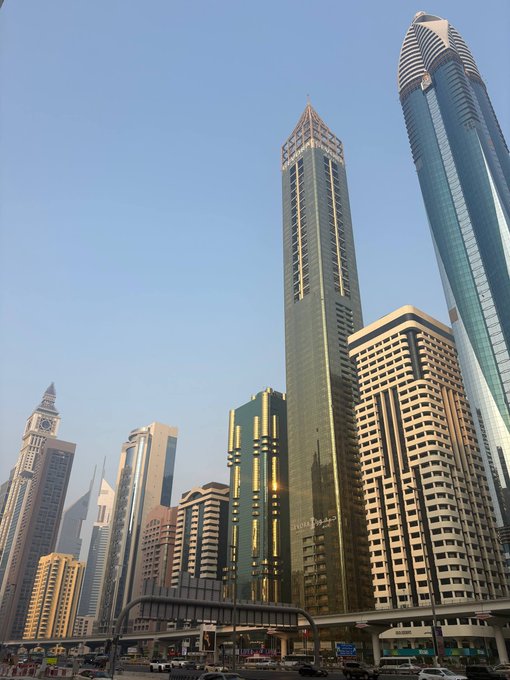 A street-level photo of Dubai's Sheikh Zayed Road skyline featuring the Emirates Towers with cars and a metro station in the foreground.