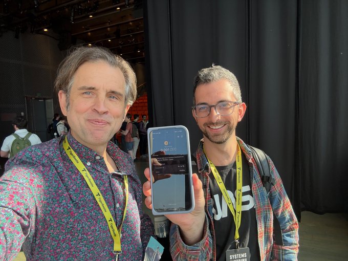 Two smiling men pose for a selfie at what appears to be a tech conference, with one proudly displaying a smartphone showing the Zcash logo on screen, celebrating Jim's new status as a "Zcasher." Both wear lanyards and the man on the right has a badge labeled "SYSTEMS," suggesting their connection to the Bluesky and Zcash communities mentioned in the post.