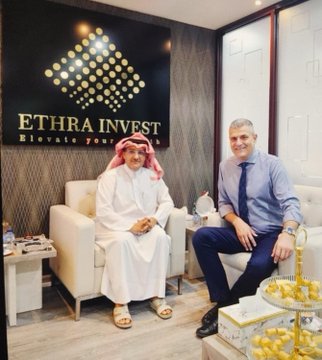 Two men—one in traditional Saudi attire and another in a blue shirt and tie—sit together in a lounge area at the **ETHRA INVEST** office (with the tagline "Elevate your..." visible on the wall behind them), posing beside a decorative tower of dates in what appears to be a business meeting setting related to the Ethra shipping/investment venture.