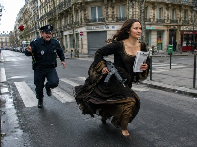 A woman in a flowing brown dress running down a city street holding an assault rifle and a newspaper, while a uniformed police officer chases her.